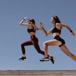 Two women in active wear leap energetically on stairs against a clear blue sky.