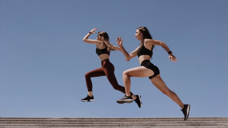 Two women in active wear leap energetically on stairs against a clear blue sky.